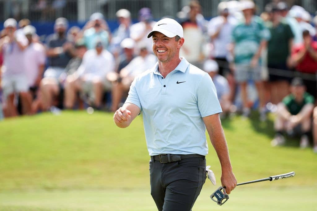 Rory McIlroy smiles as he walks past a blurred crowd on the green, swinging his right arm forward and holding a club in his left foot. He wears grey jeans with a glove poking out from his lefthand back pocket, a pastel blue polo with a dark Nike logo and a white cap with a dark Nike logo.