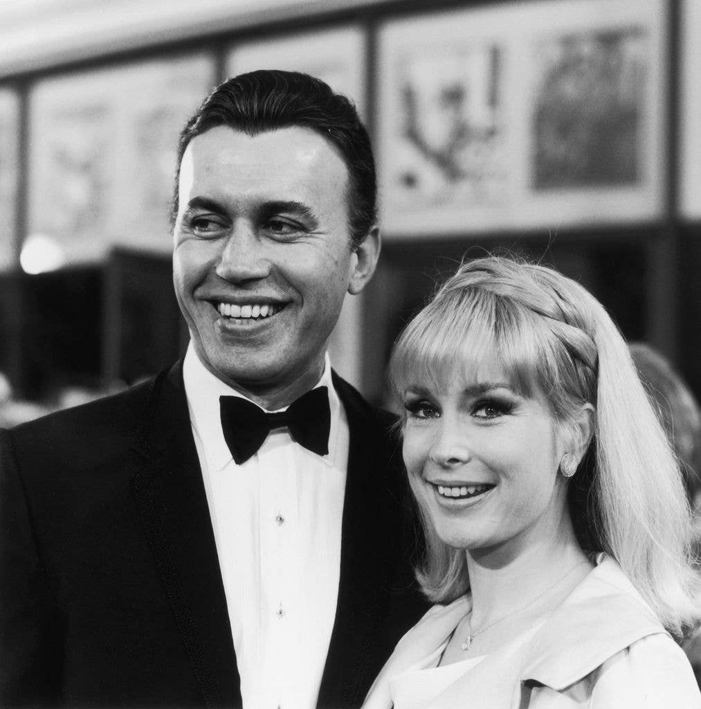 Black and white photo of Michael Ansara in a bowtie and suit standing next to wife Barbara Eden who holds a section of hair back with a braid. 