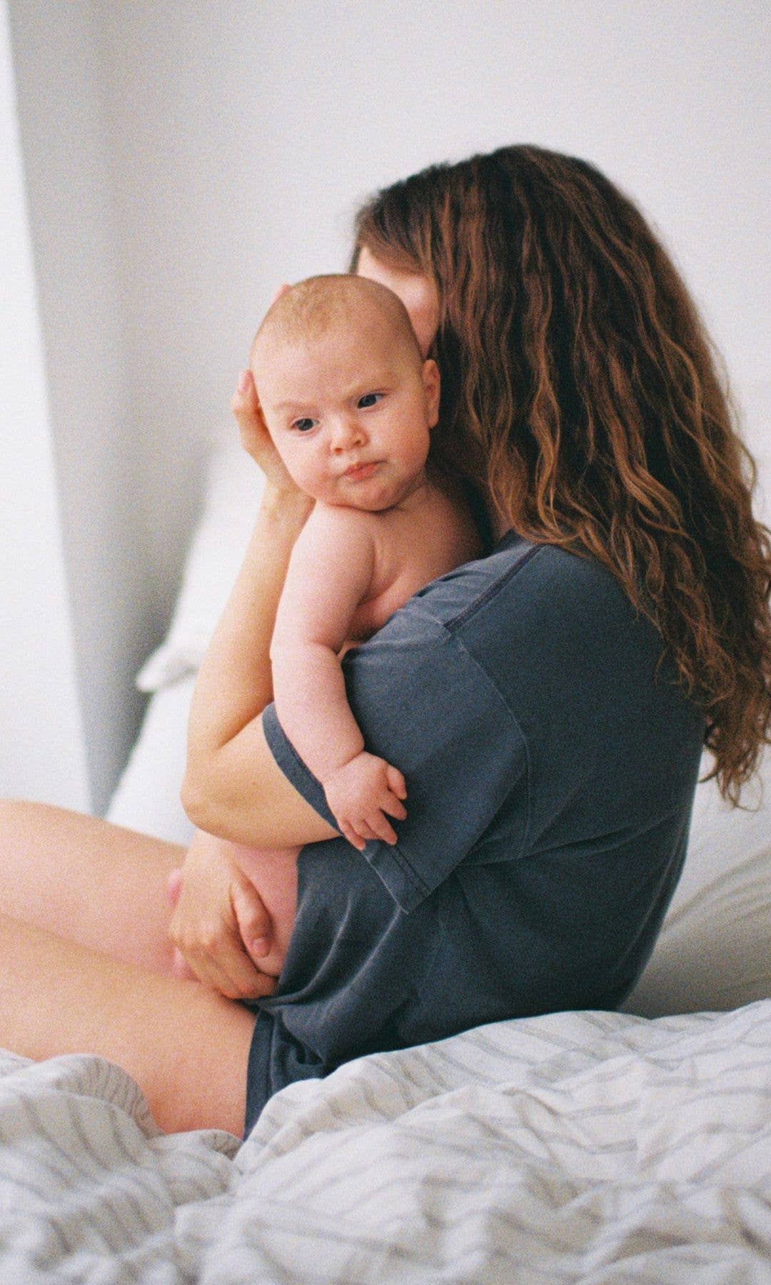 A woman in a grey t-short on a bed holding a baby