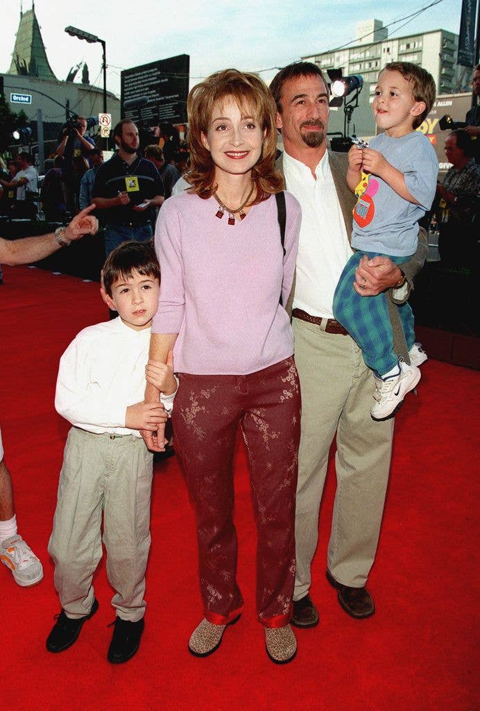 Annie Potts, husband James Hayman and sons Harry and Doc stand on the red carpet. Both James and son Harry wear khaki trousers with a white shirt, while Annie holds onto son Harry and James' hands wearing a pink jumper with red patterned trousers. James holds son Doc in one arm, who wears checked teal trousers and a grey graphic tee.