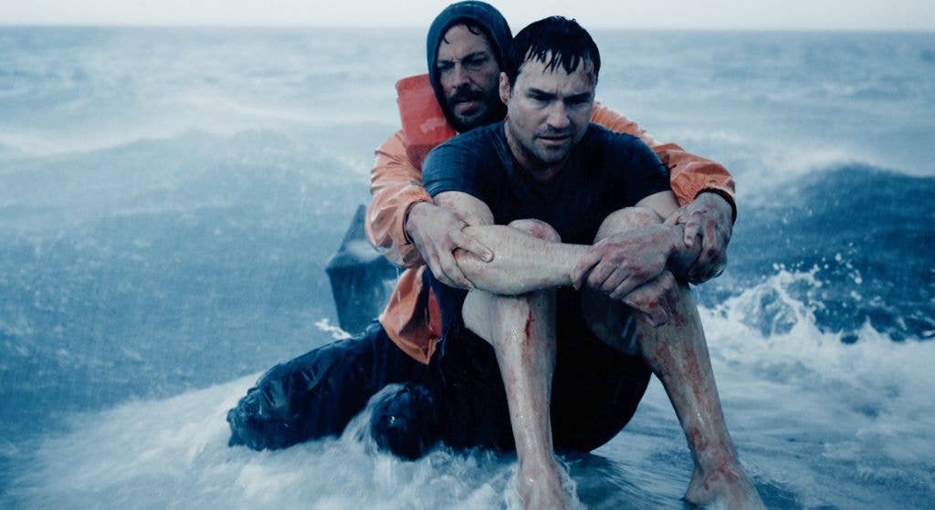 A man with short hair is being supported by another person in an orange life jacket in rough ocean waters.