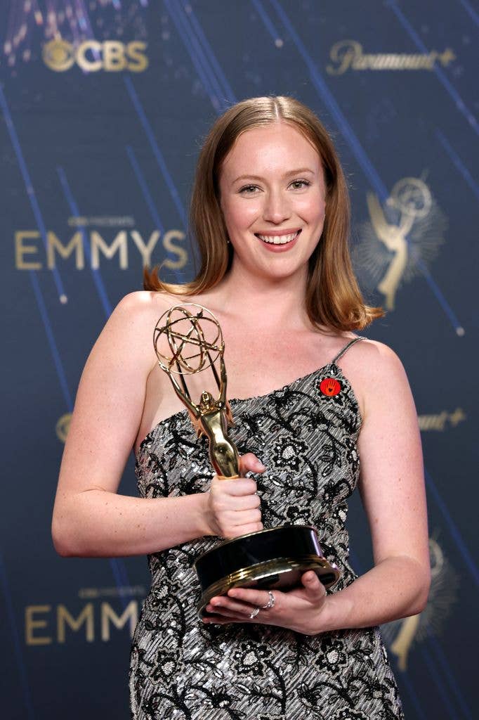 Hannah Einbinder wears a sparkly black and grey, one strap dress with a memorial poppy affixed. She holds an Emmy award in two hands.