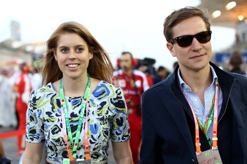 Princess Beatrice and David Clark walk across the grid during the Bahrain Formula One Grand Prix in 2015. Beatrice wears a short sleeved white dress with a blue, black and green pattern while then boyfriend Dave wears a striped blue shirt and half zipped jacket.