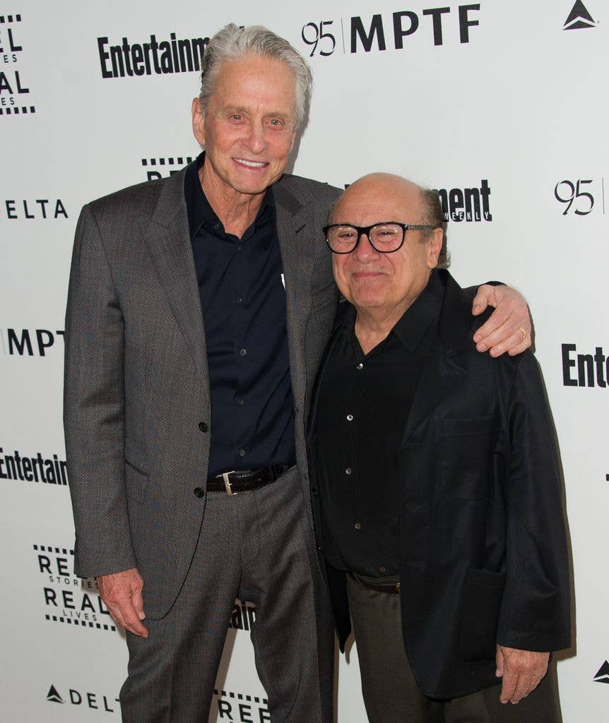 Michael Douglas in a grey suit with a black shirt stands next to Danny Devito in dark grey trousers, a black shirt and a black jacket.