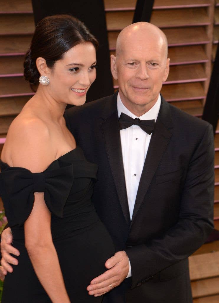 Bruce Willis holds a hand on wife Emma Hemming's stomach. Bruce wears a tux and black bowtie while Emma wears an off the shoulder black dress.