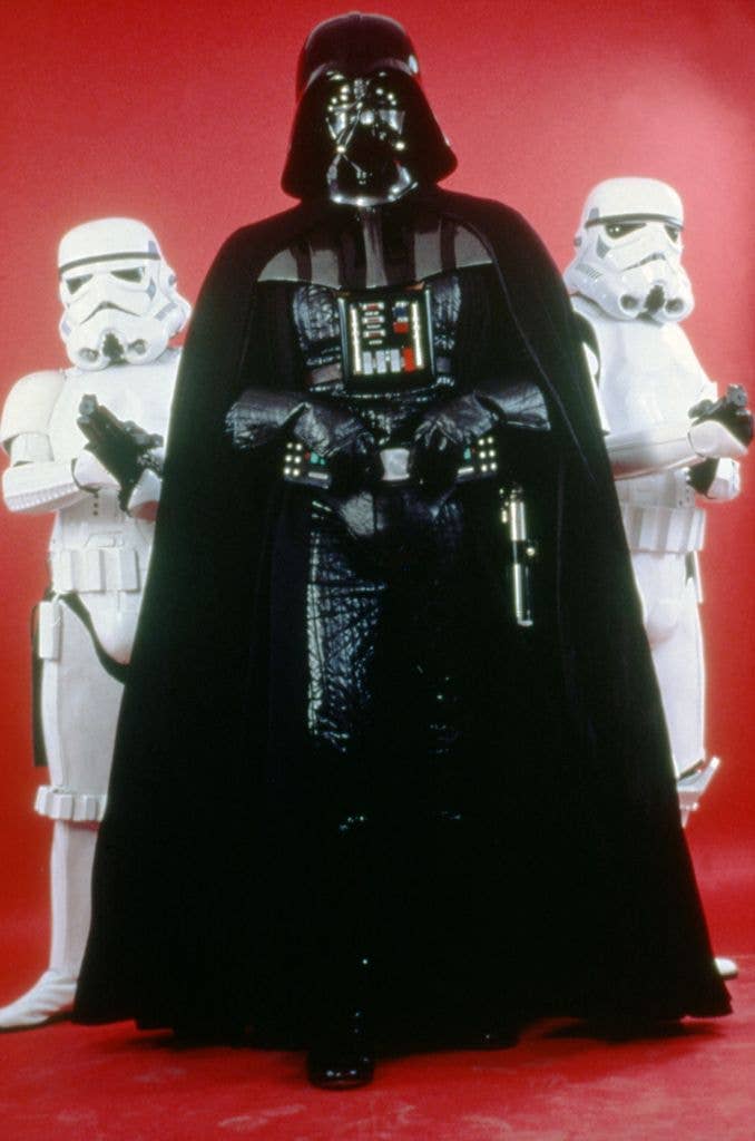 British actor David Prowse on the set of Star Wars: Episode V - The Empire Strikes Back poses with two stormtroopers in front of a red background.
