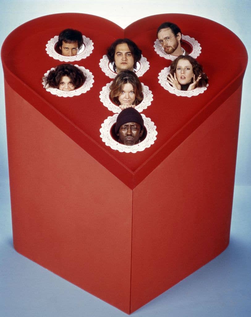 Chevy Chase, John Belushi, Michael O'Donoghue, Gilda Radner, Jane Curtin, Laraine Newman, and Garrett Morris all peek out from a large heart shaped box.