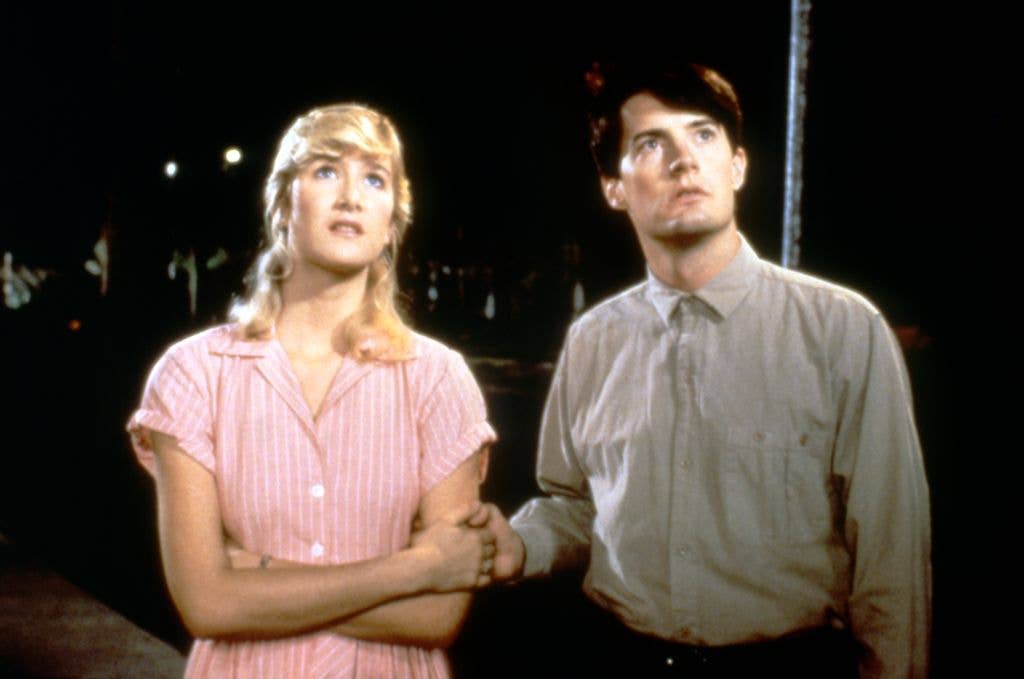On the set of 'Blue Velvet', Laura Dern wears a pink and white stripe dress, folding her arms, while Kyle MacLachlan wears a grey shirt and holds her hand. Both look up towards the top of the image.