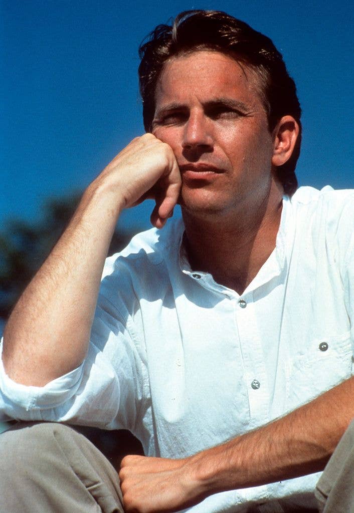 Kevin Costner in a scene from the film 'Field Of Dreams' wearing a white shirt and grey trousers, resting his elbow on his right knee and his face on his knuckles.