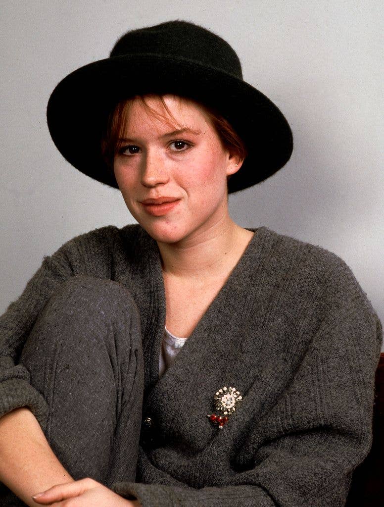 Portrait of Molly Ringwald in a dark grey cardigan and dark grey trousers, pulling her knee up to her chest with clasped hands, wearing a dark wide brim hat.