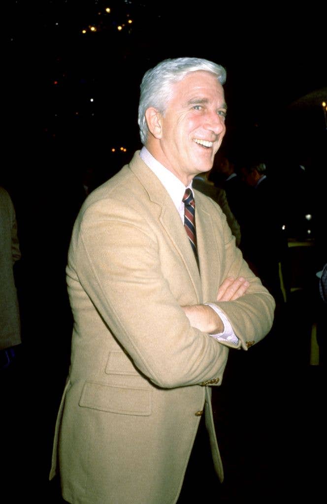 Leslie Nielsen in a tan suit with a white shirt and striped tie crosses his arms and smiles.