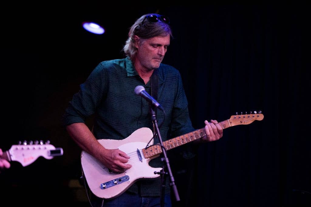 Val McCallum performs on stage with a guitar, wearing a dark shirt and looking down at the instrument.
