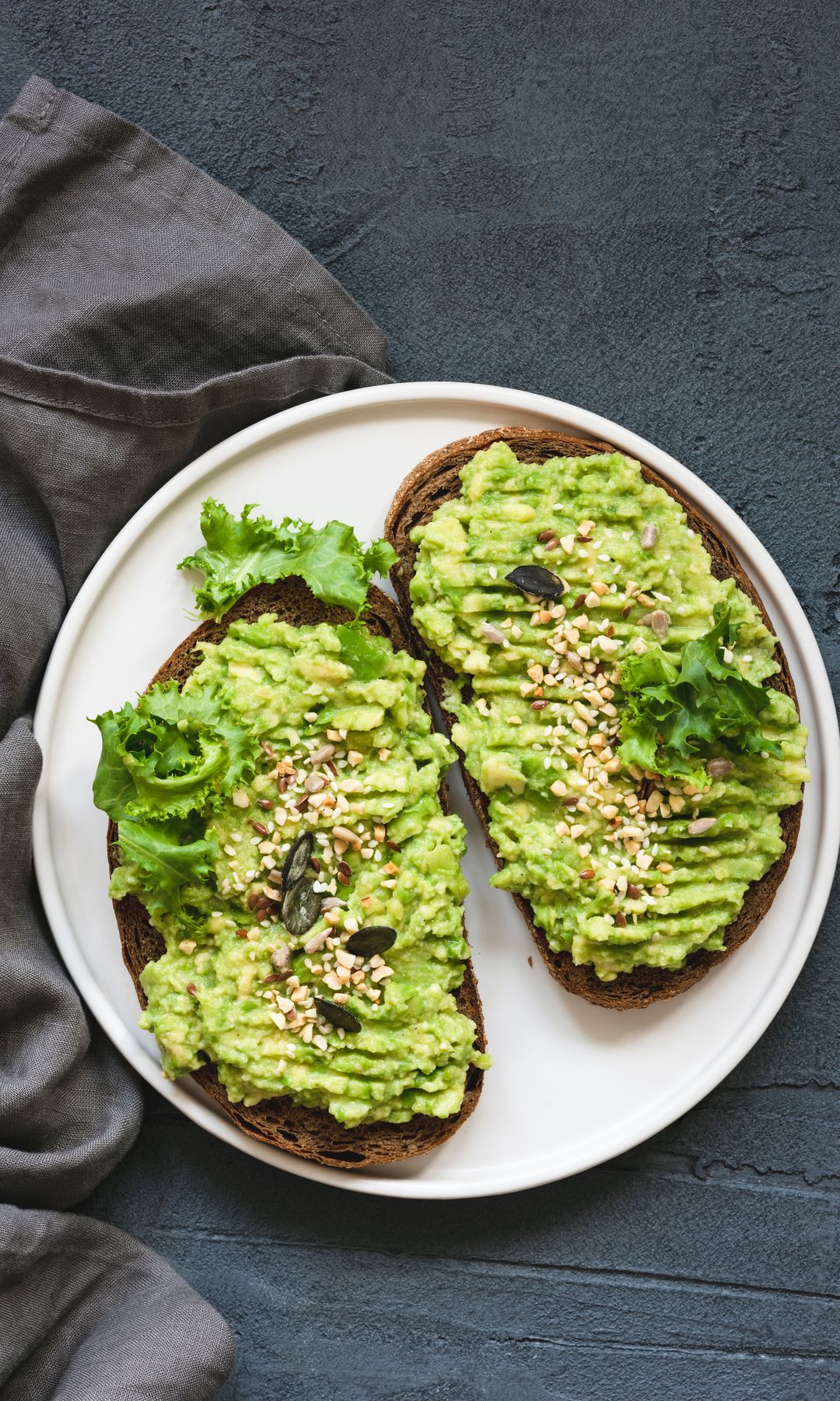 Two rye toasts with mashed avocado, seeds and green salad on white plate. Top view