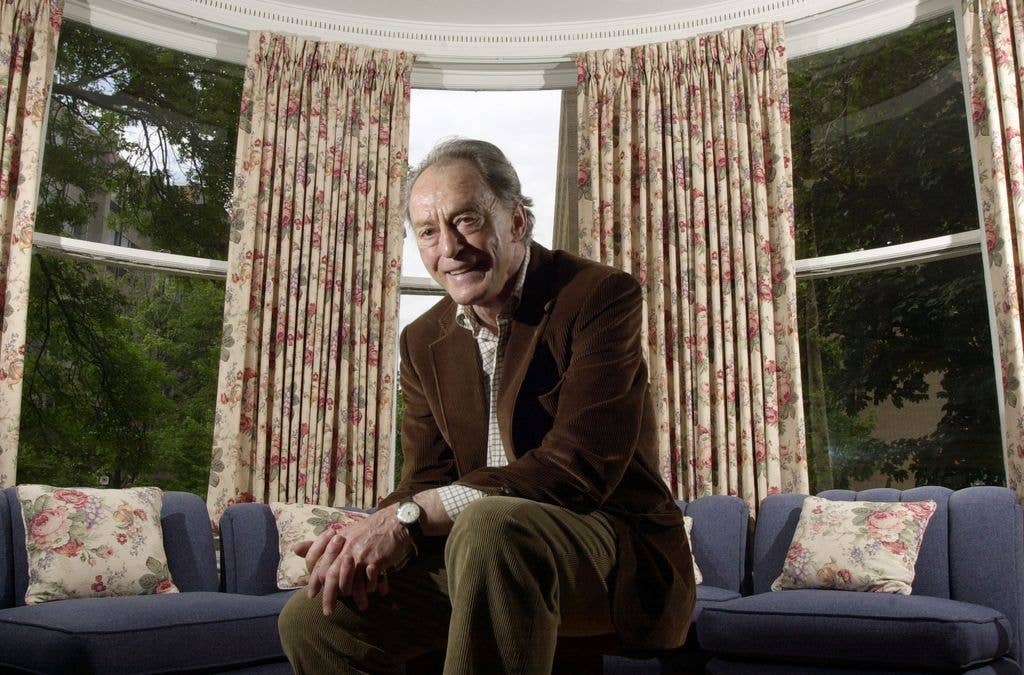 Peter Donat sits in front of a blue sofa in a curved window , wearing a brown jacket and khaki trousers.
