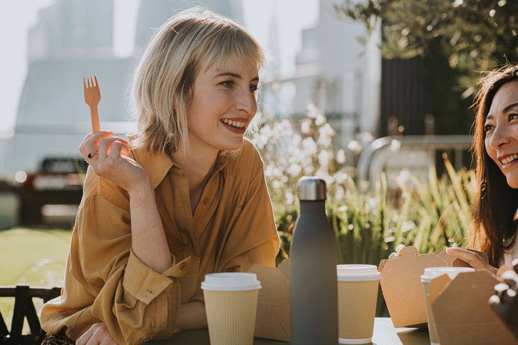 A woman smiles as she enjoys an al fresco lunch with another woman. She holds a wooden fork, and eats from takeout containers.