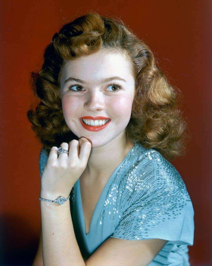 Shirley Temple in a blue dress, holding her chin in her left hand and looking to the right of the image.