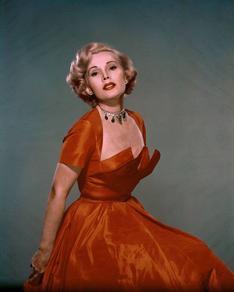 Zsa Zsa Gabor in an orange satin dress posing against a grey background.