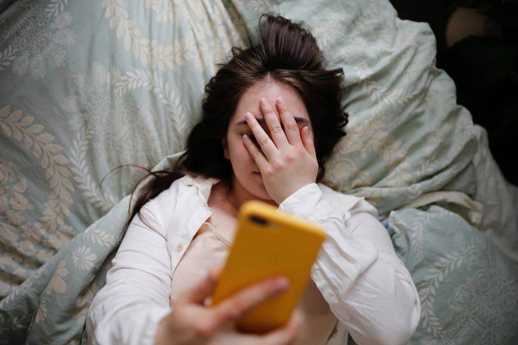 A woman in a white shirt lies in bed, covering her face with her hand while holding a yellow smartphone.