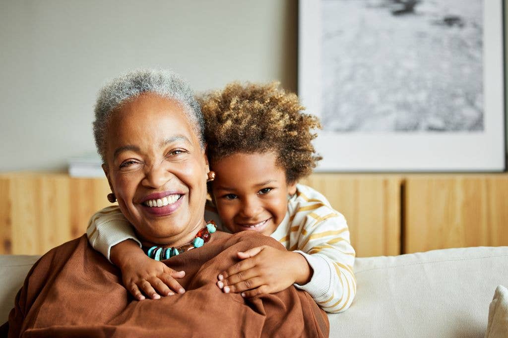 woman with grey hair, brown top, brown earrings and turquoise and brown necklace is hugged from behind by her grandchild, wearing a striped shirt