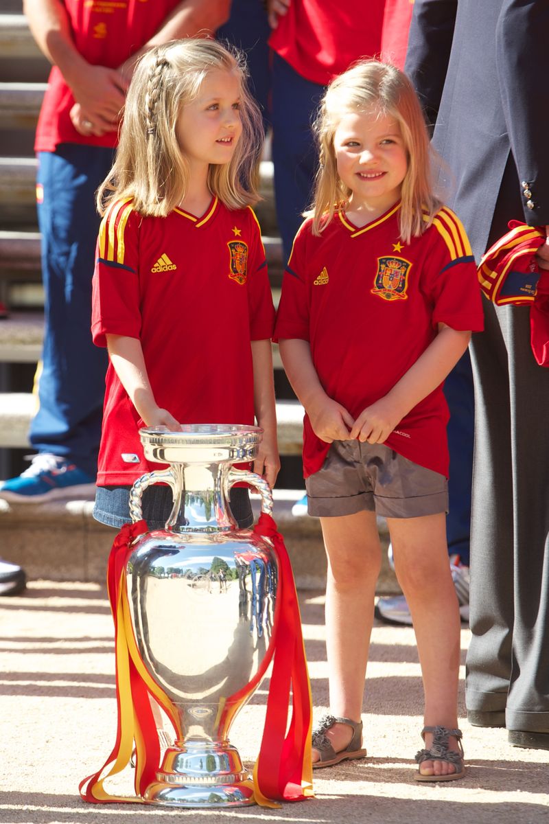 MADRID, SPAIN - JULY 02: Princess Leonor of Spain (L) and Princess Sofia of Spain stand next to the UEFA EURO 2012 trophy as King Juan Carlos I of Spain receives players of Spain's victorious national football team at Zarzuela Palace on July 2, 2012 in Madrid, Spain. (Photo by Pool/Getty Images)