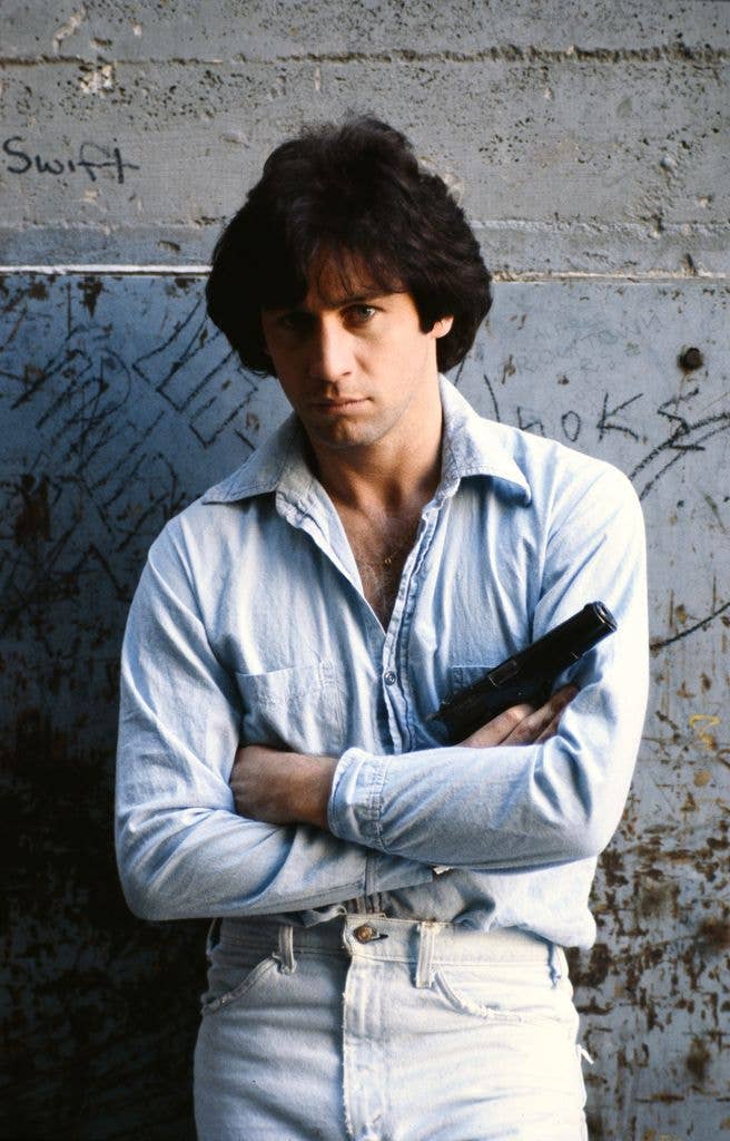 Joseph Travolta stands cross armed against a wall in a blue shirt and white jeans.