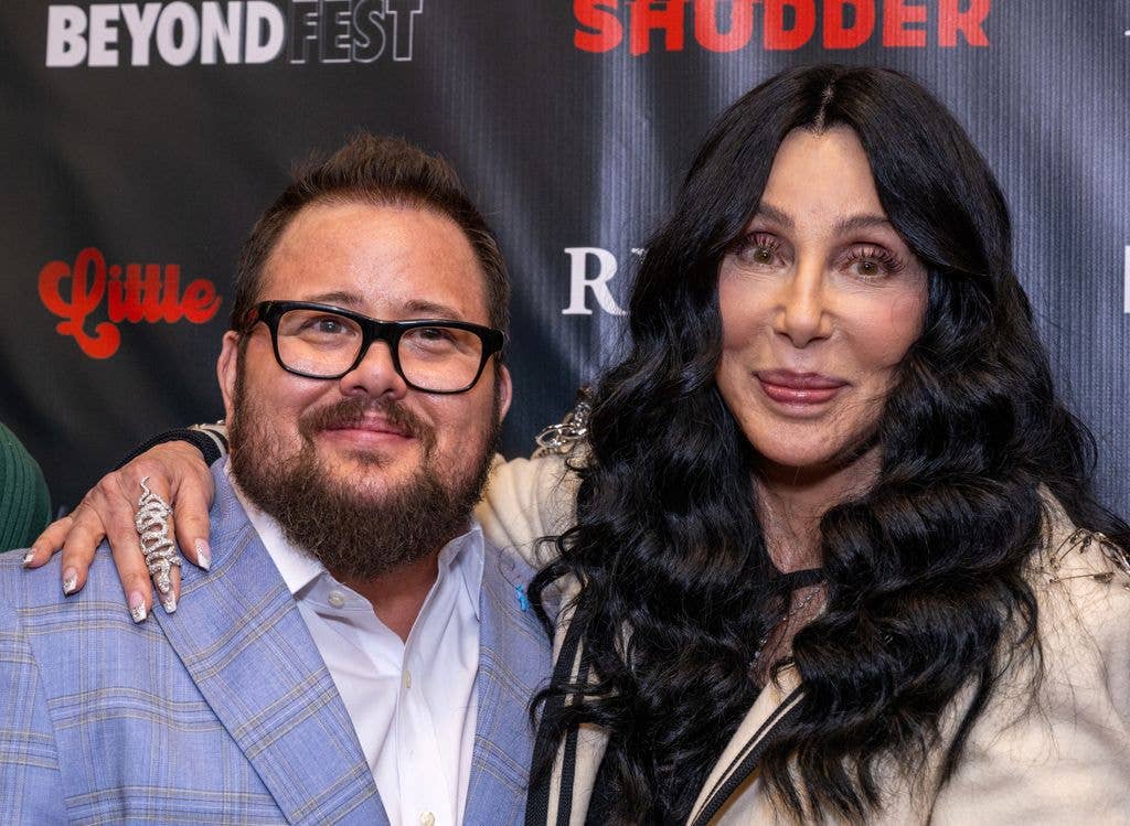 Chaz Bono in a blue suit and white shirt, standing next to his mother Cher who holds an arm around her son.