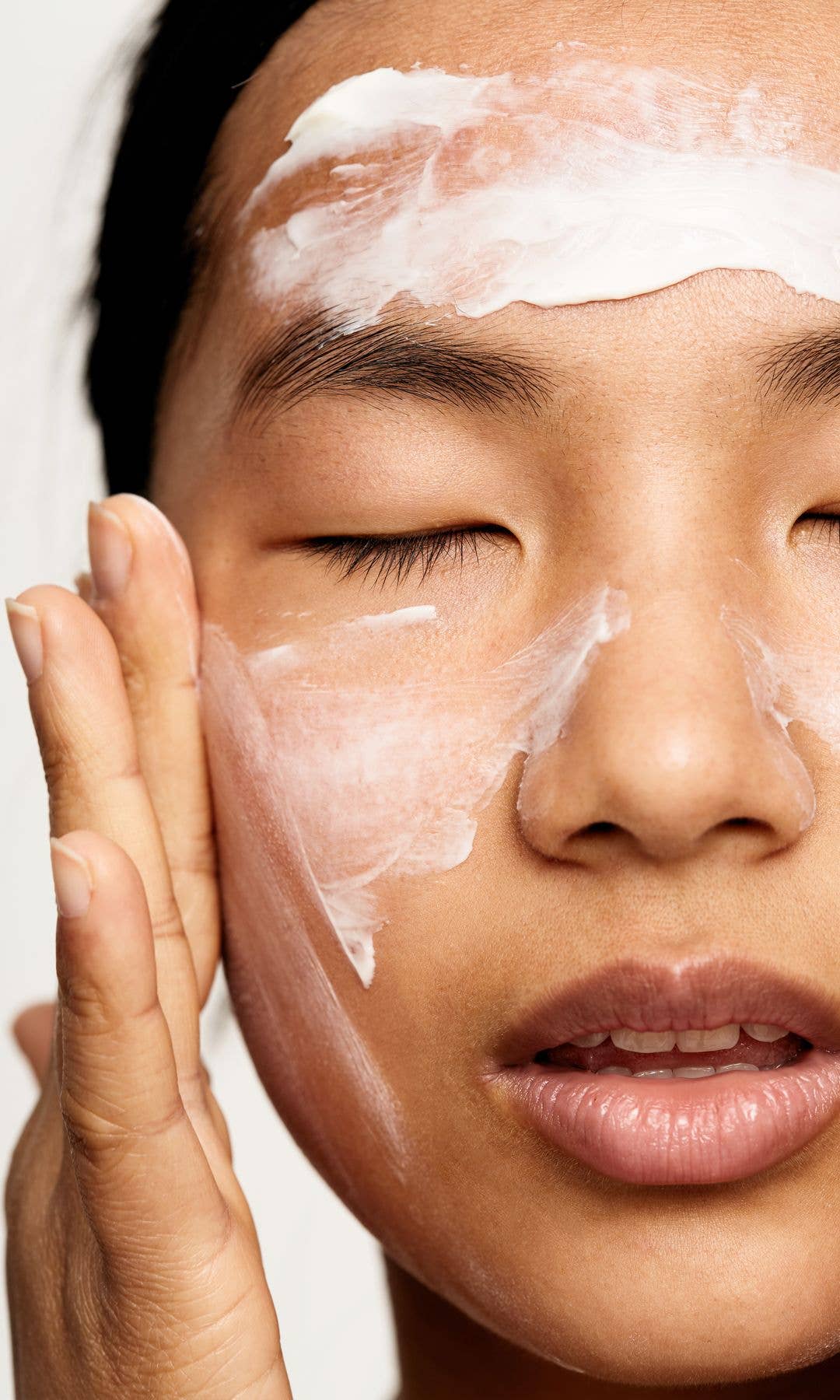 Asian model with her eyes closed applying a white skincare cream