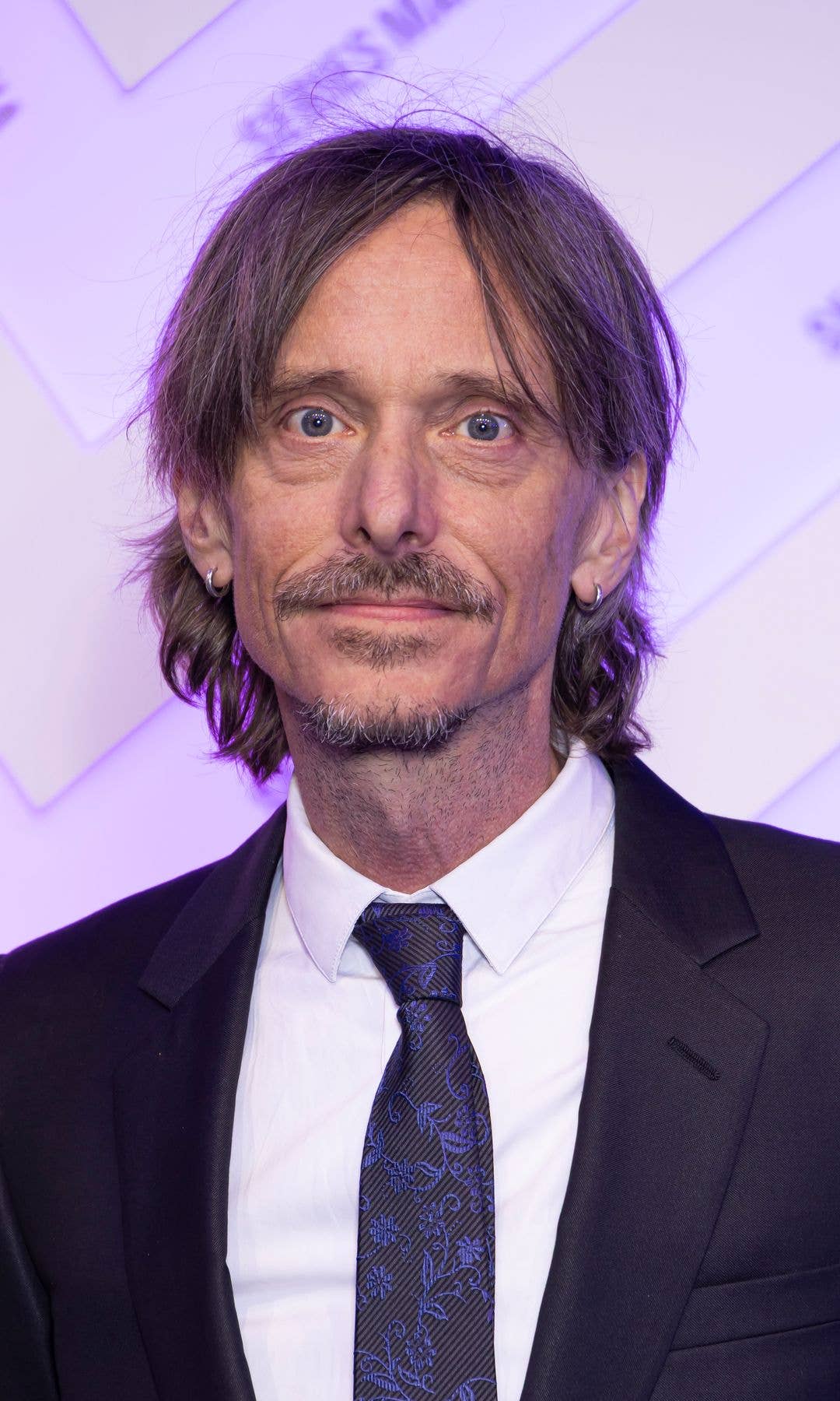 Mackenzie Crook in a black suit and blue tie, smiling at the camera.