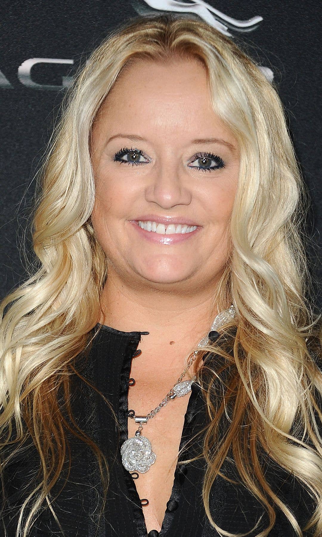 Lucy Davis smiling, wearing a black top and a silver necklace with a flower pendant.