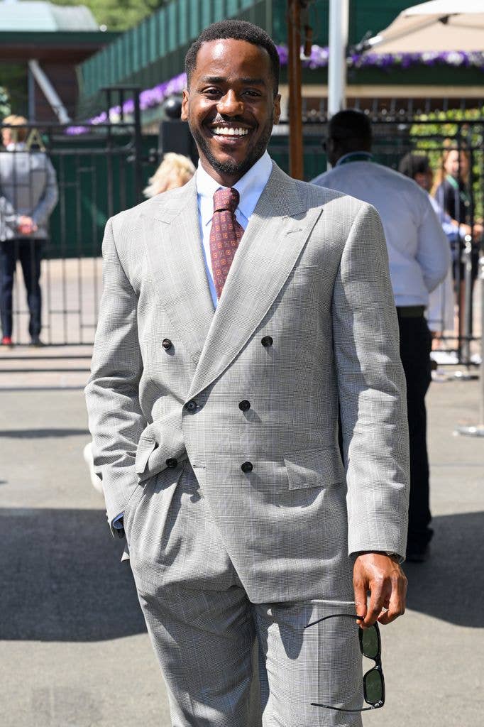 Ncuti Gatwa in a gray double-breasted suit, red tie, and green sunglasses, smiling.