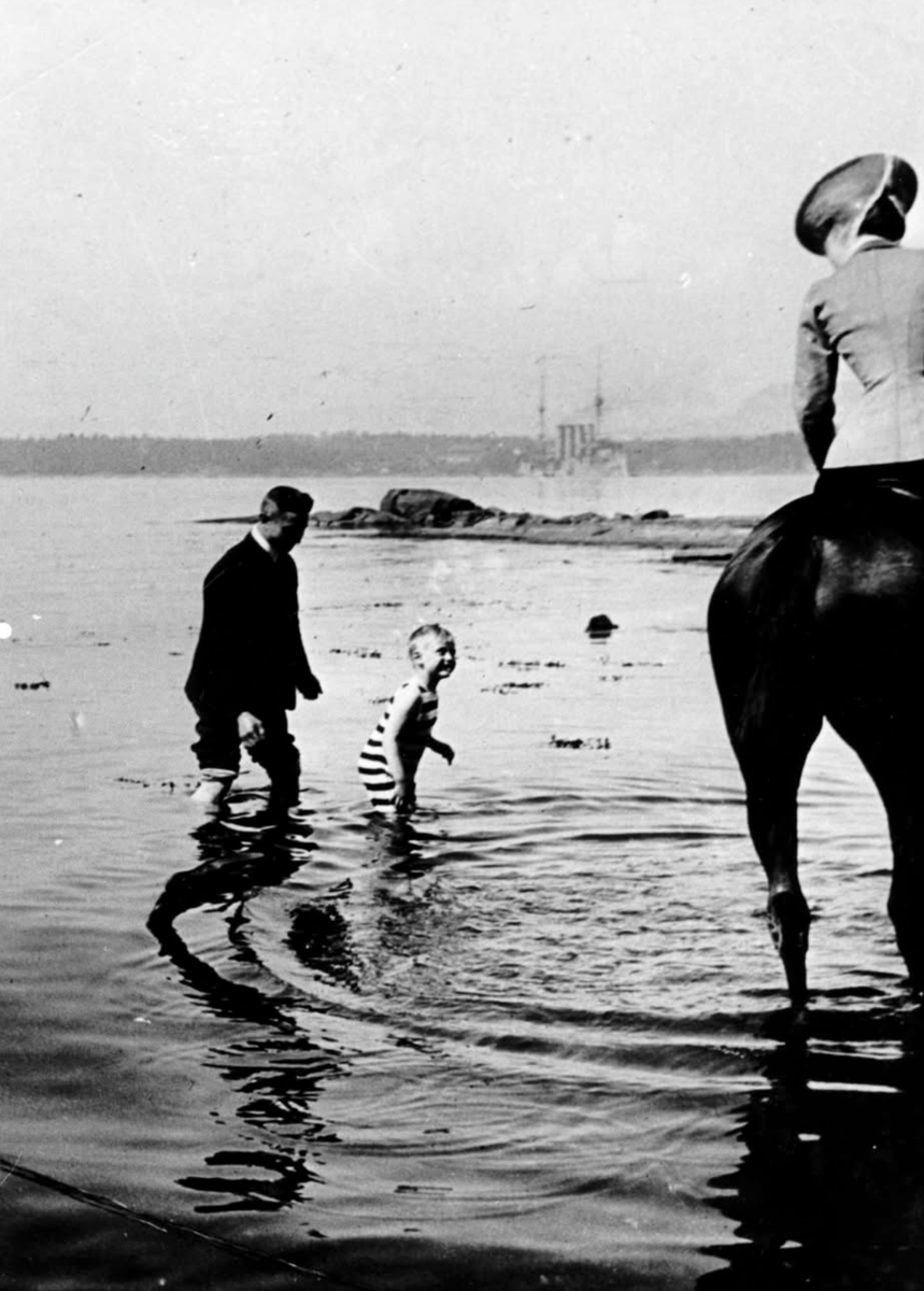 Queen Maud rides on horseback while Crown Prince Olav plays in the water in 1910.