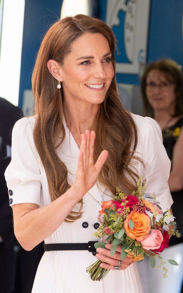 Kate Middleton in a white dress, holding a colorful bouquet, waving.