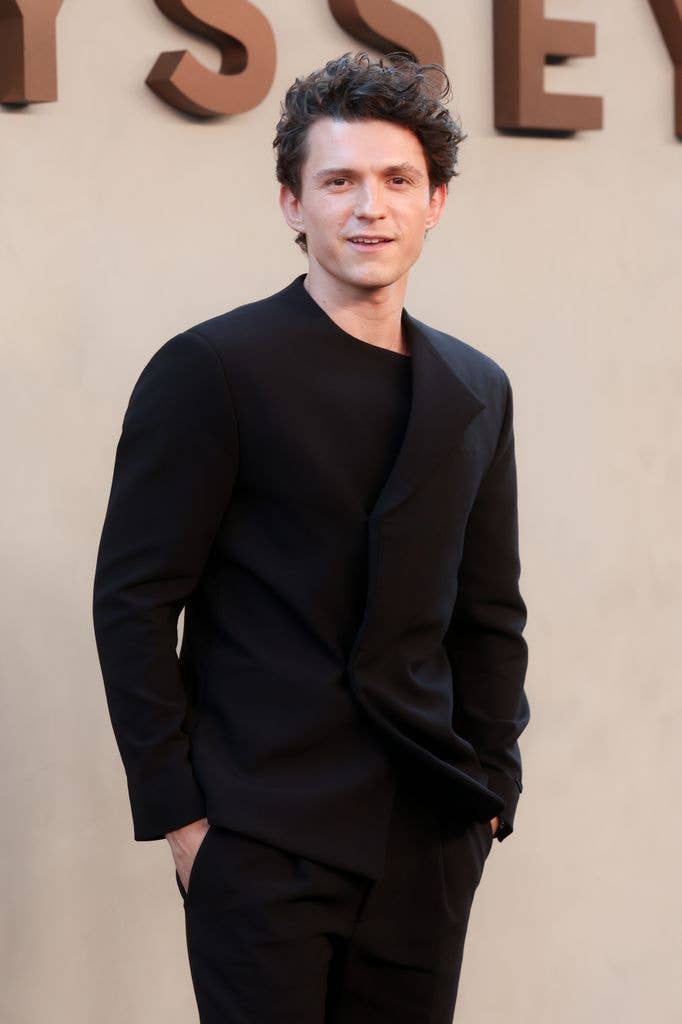 Tom Holland stands in front of a wall with large letters, wearing a black suit.