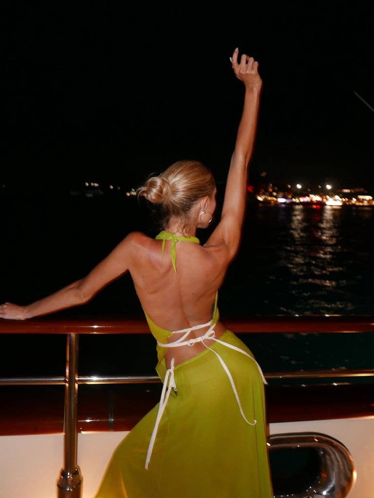 Woman in a green dress stands on a boat deck at night, arm raised.