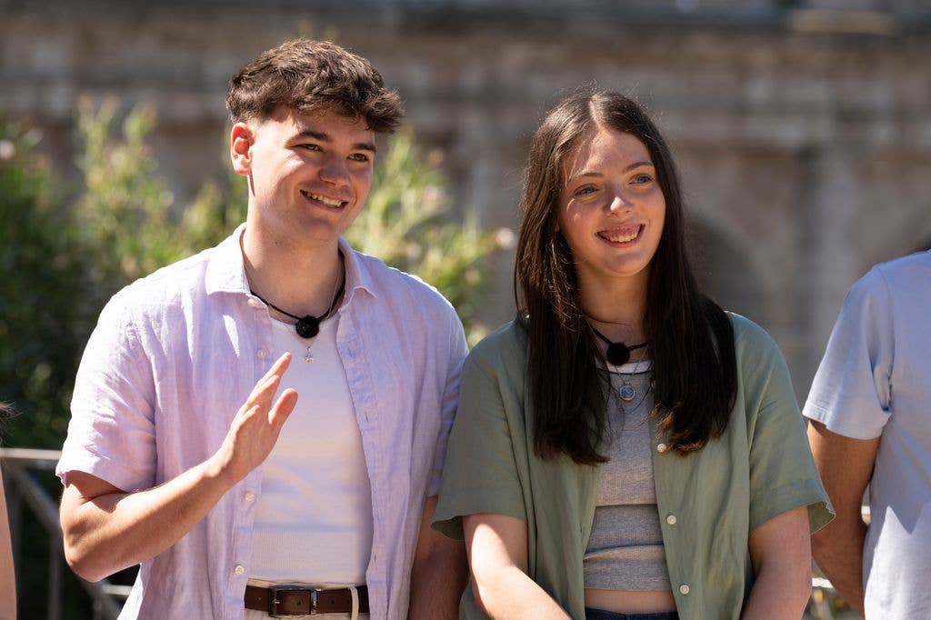 Two smiling individuals stand close together, one in a white shirt and the other in a green jacket.