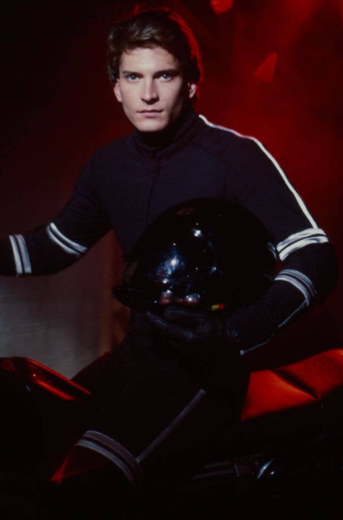 Rex Smith in a black outfit with white stripes, holding a helmet, seated on a motorcycle.