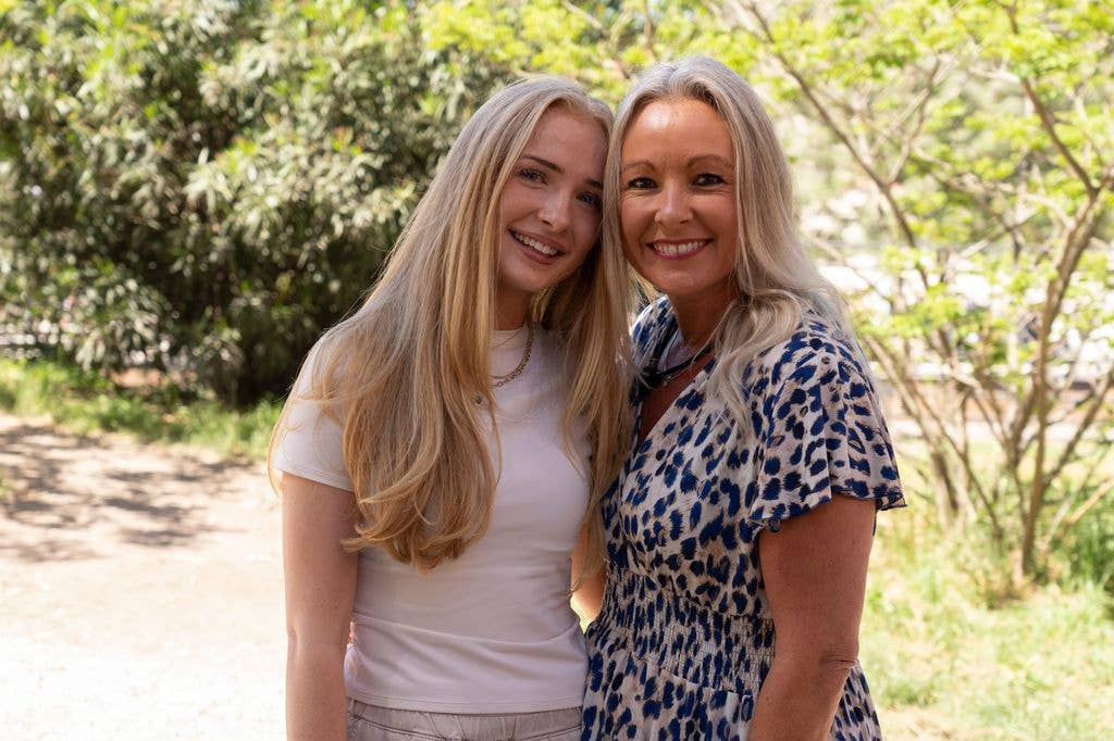 Two women with blonde hair stand together, smiling, in a park setting.
