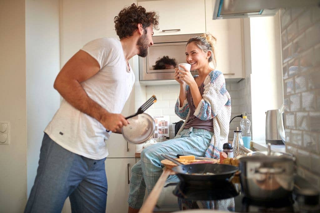 Man in white t-shirt and jeans holding a pan, talking to a woman in a cardigan and jeans holding a cup.