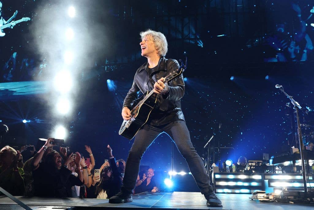 Jon Bon Jovi with white hair and black leather jacket playing guitar on stage with crowd cheering.
