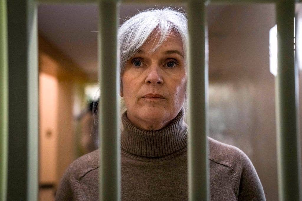 Siobhan Finneran stands behind bars, wearing a brown turtleneck sweater.