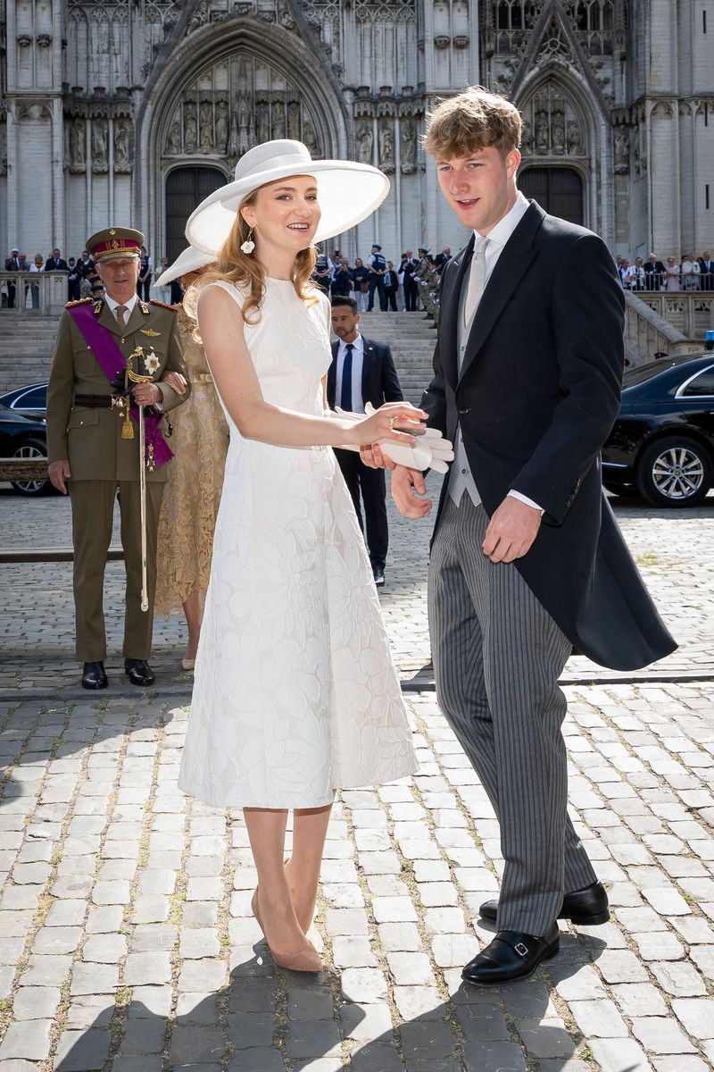 Princess Elisabeth of Belgium, in a white dress and wide-brimmed hat, walks arm in arm with Prince Emmanuel in a morning coat outside a Gothic cathedral