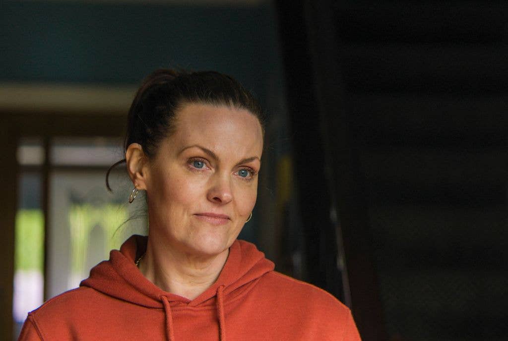 Jo Joyner in a red hoodie, standing near a doorway with a serious expression.