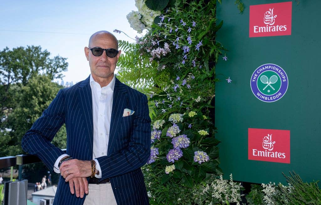 Stanley Tucci stands in a blue pinstripe suit, white shirt, and sunglasses, next to floral decor and a Wimbledon sign.