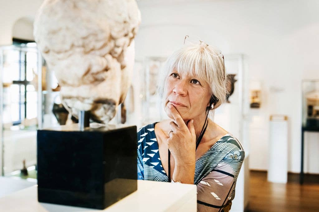 A woman with short blonde hair stands by a marble bust, hand on chin, in a museum.