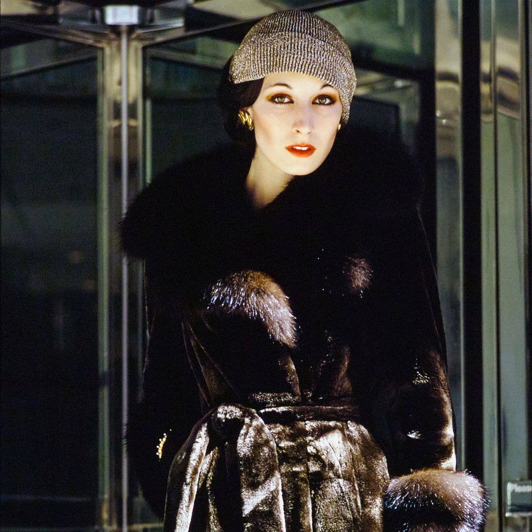 Model and actress Anjelica Huston standing in front of a revolving door wearing sealskin wrap-and-tie fur coat with sable collar and cuffs by Emeric Partos. Made of Fouke-processed silver Safari Cape fur seal. Lurex ribbed knit cap by Don Anderson for Scheer Brothers, Aris Gloves, and jewels from Tiffany & Co. Hair by William of Kenneth. 