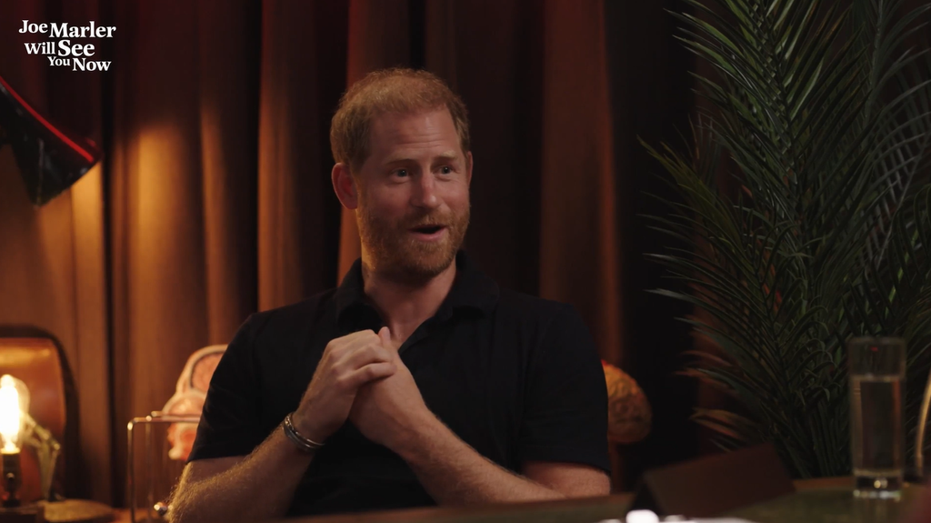 Prince Harry in a black shirt, sits at a table with his hands clasped
