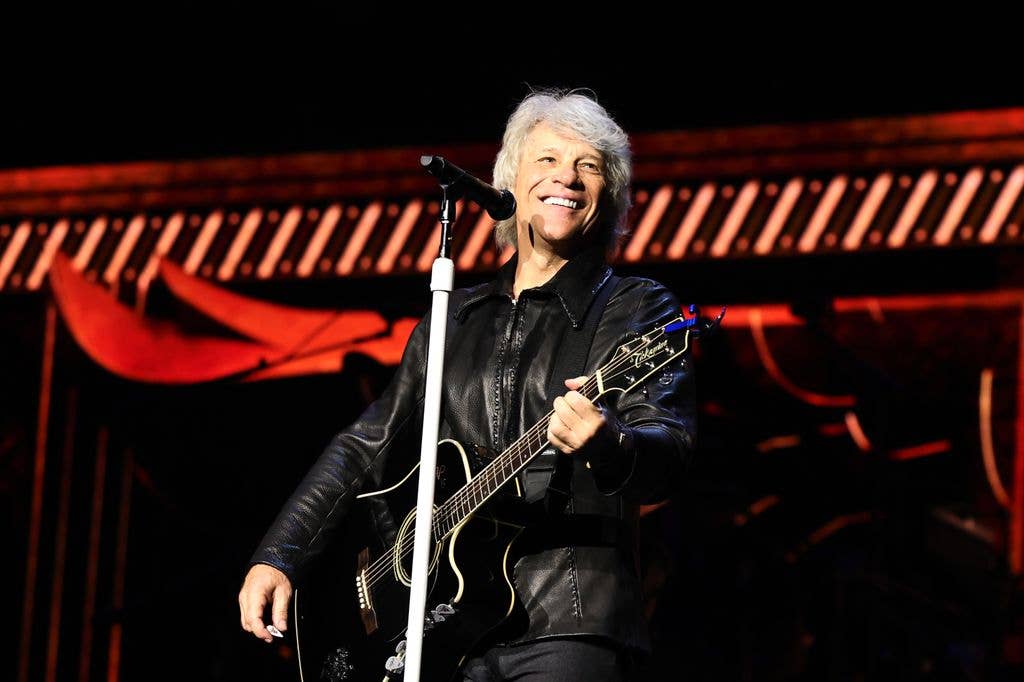 Jon Bon Jovi with white hair and black leather jacket playing guitar on stage.