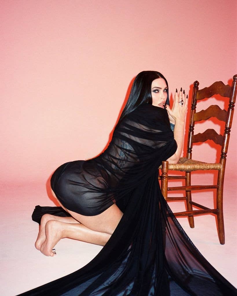 Megan Fox in a black sheer dress, kneeling next to a wooden chair.