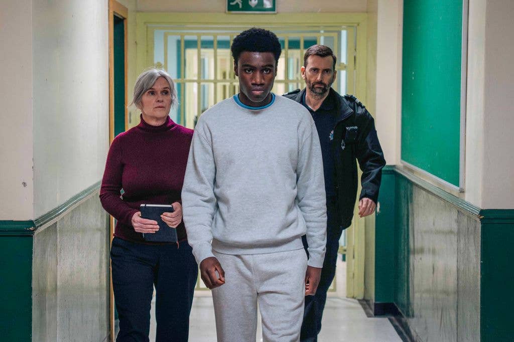 A man in gray sweats is flanked by a woman (Siobhan Finneran) in a maroon turtleneck and a man in a black uniform (David Tennant)
