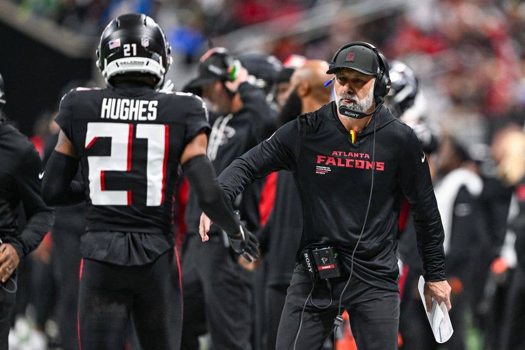 Atlanta Falcons coach Jeff Ulbrich, wearing headset and holding clipboard, talks to player Hughes on field.
